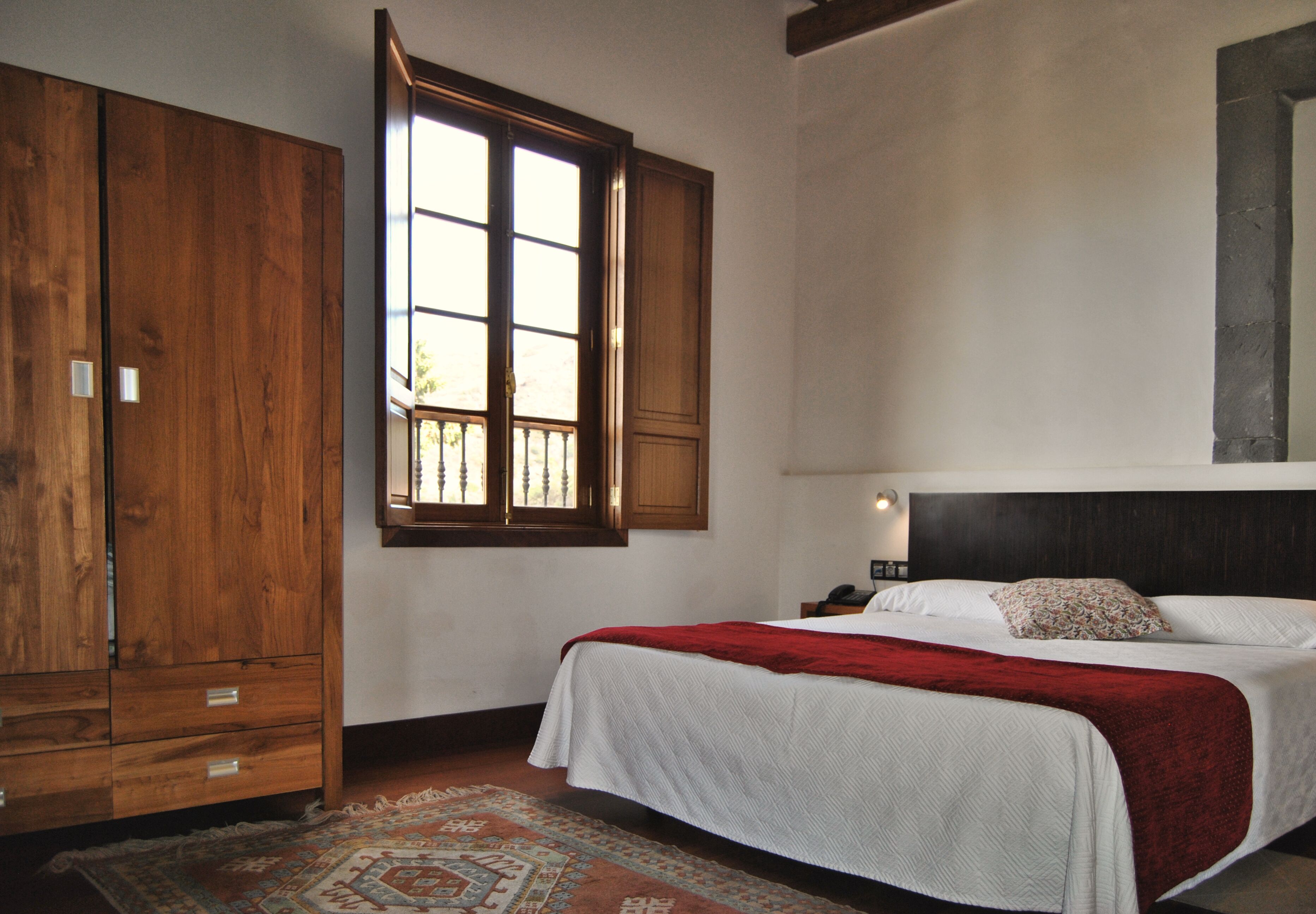 Un dormitorio con un armario de madera grande, una cama con ropa blanca y una manta roja, una mesita de noche con una lámpara y una alfombra estampada.
