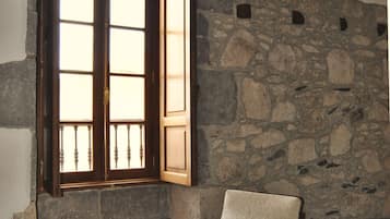 A room with a stone wall, a wooden-framed window, a cushioned chair, and a wooden floor.