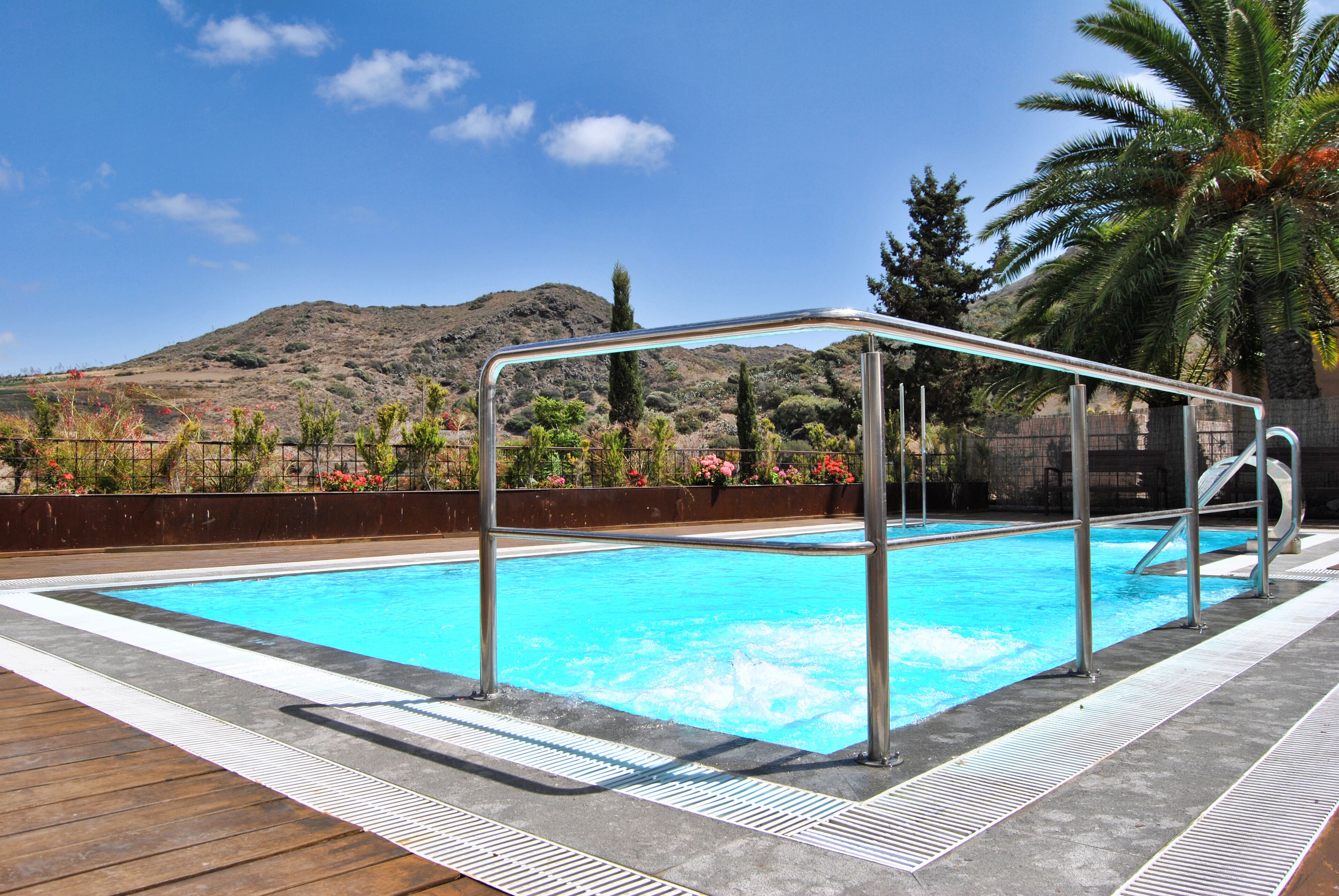 A swimming pool with a metal safety railing, surrounded by a wooden deck and a low stone wall.