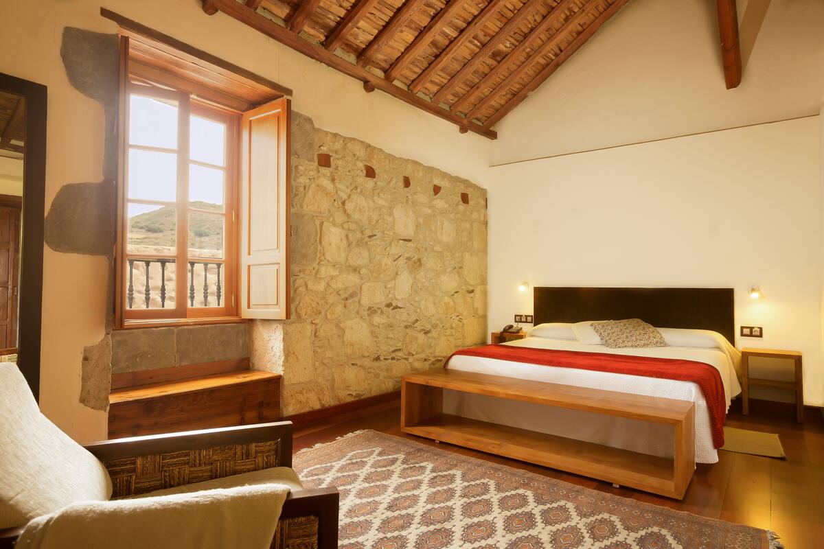 Een slaapkamer met een stenen muur, een houten bed, een nachtkastje, een bankje en een raam met balkon.