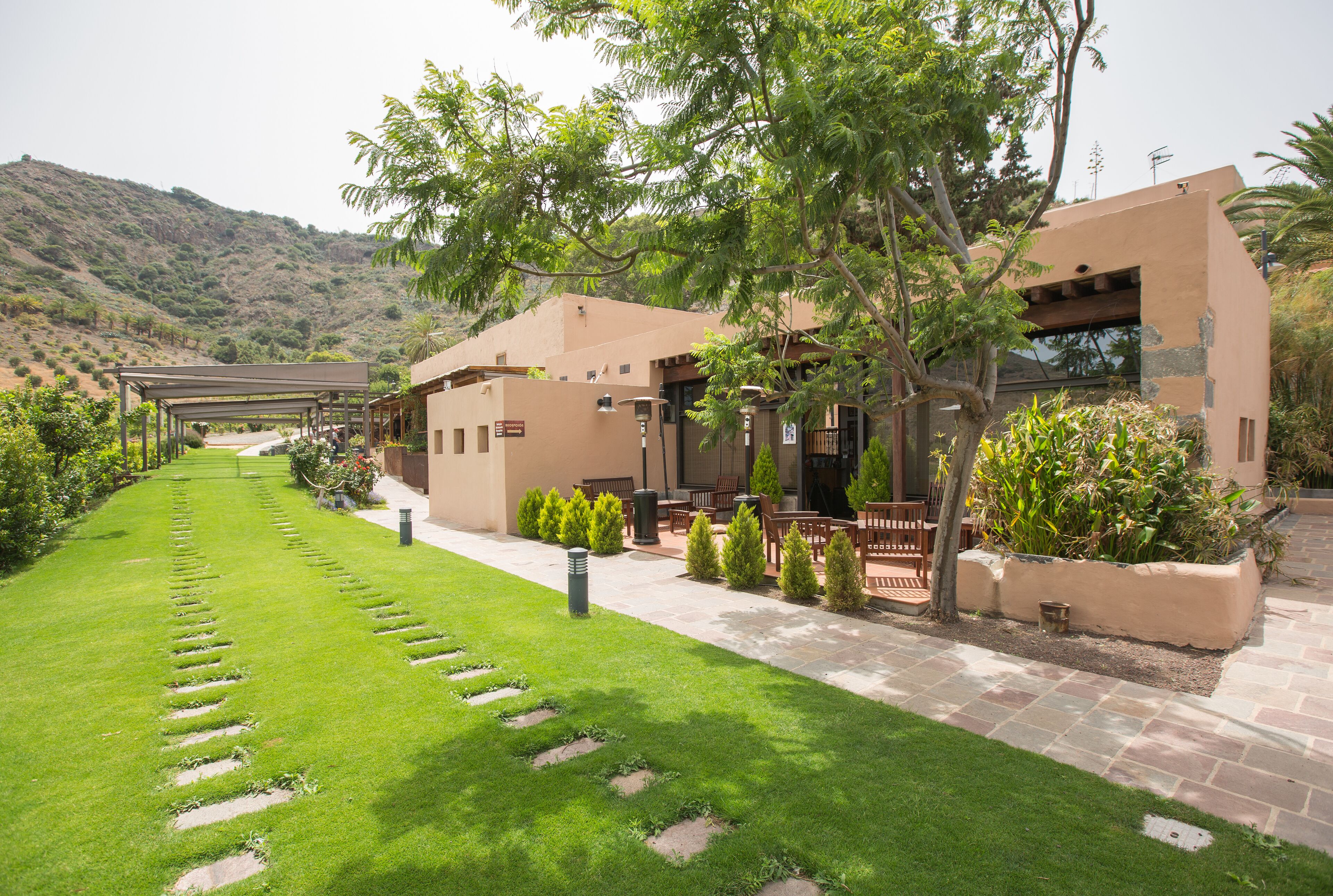 A courtyard with a paved walkway, a covered seating area, and a building with a flat roof and beige walls.