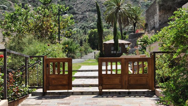A stone pathway leading to a wooden gate with a view of a hillside and a building in the background. - Hotel Rural El Mondalón (Las Palmas de Gran Canaria)