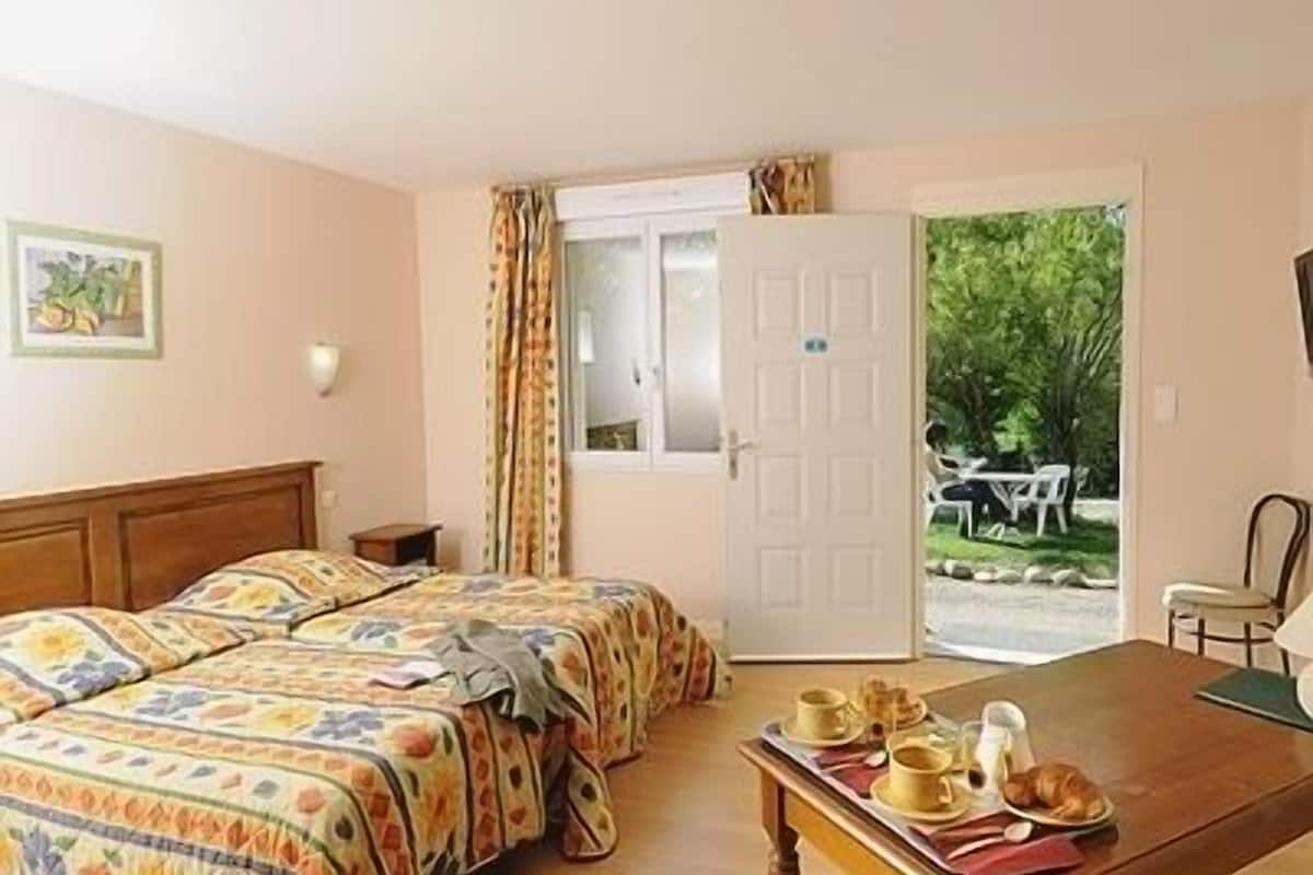 A room with two beds, a dining table with breakfast items, a window with curtains, and a view of the outdoors.