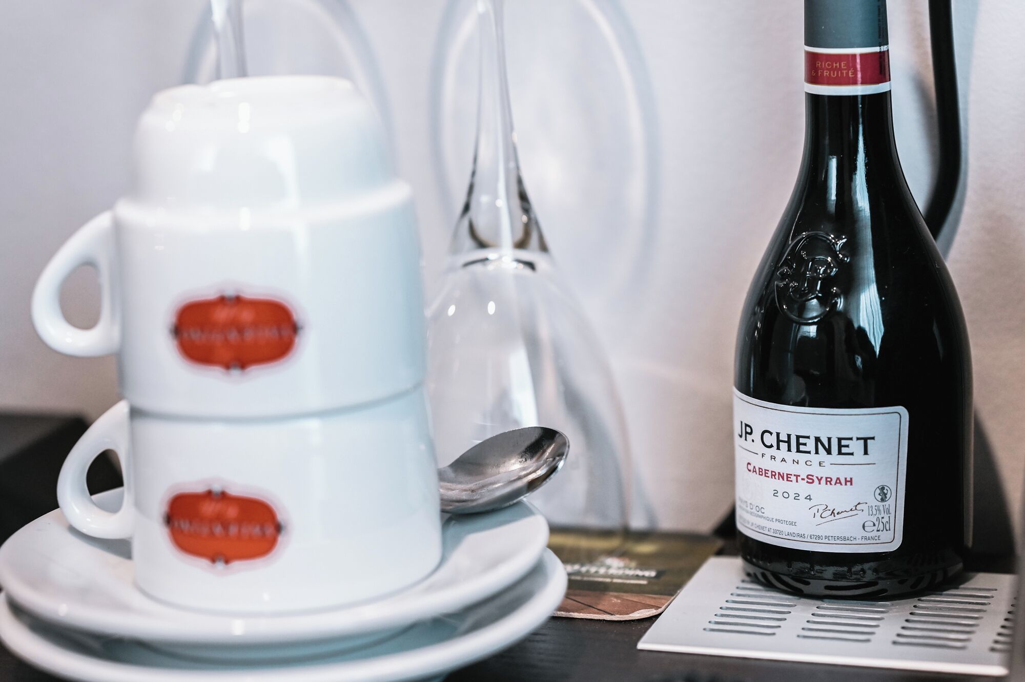 A white ceramic cup with a red design, a wine bottle labeled "JP. CHENET FRANCE CABERNET-SYRAN 2024", and a spoon on a table.