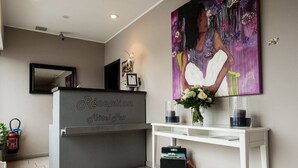 Zona de recepció d'hotel amb una taula de recepció identificada com a “Réception Hôtel Paxi”, un gran quadre floral a la paret, una taula consola blanca amb un ram de flors i un extintor a l'esquerra.