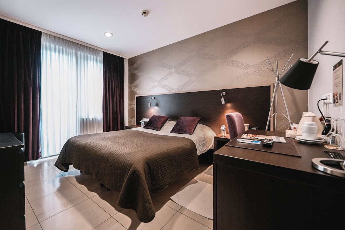A modern hotel room with a large bed, a desk with a lamp, and a view of the city through the window.