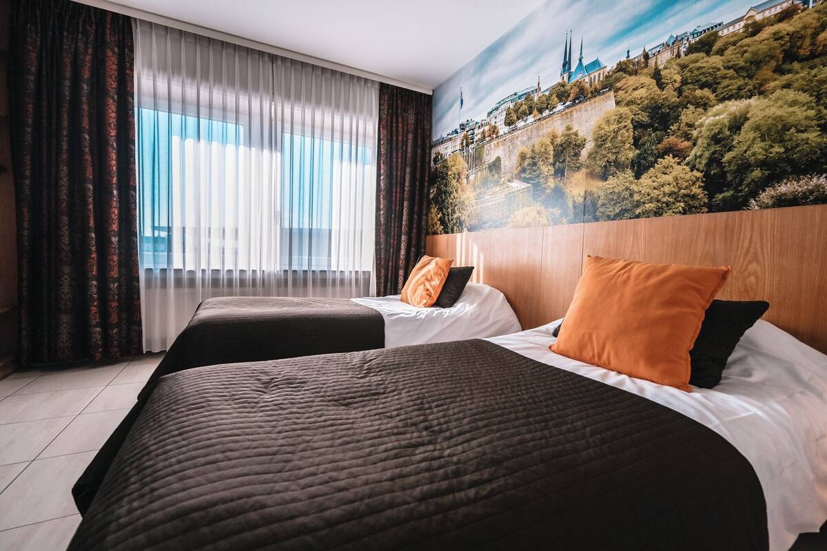 A hotel room with two beds, a large window with sheer curtains, and a mural of a cityscape on the wall.