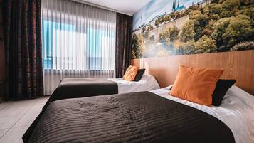 A hotel room with two beds, a large window with sheer curtains, and a mural of a cityscape on the wall.