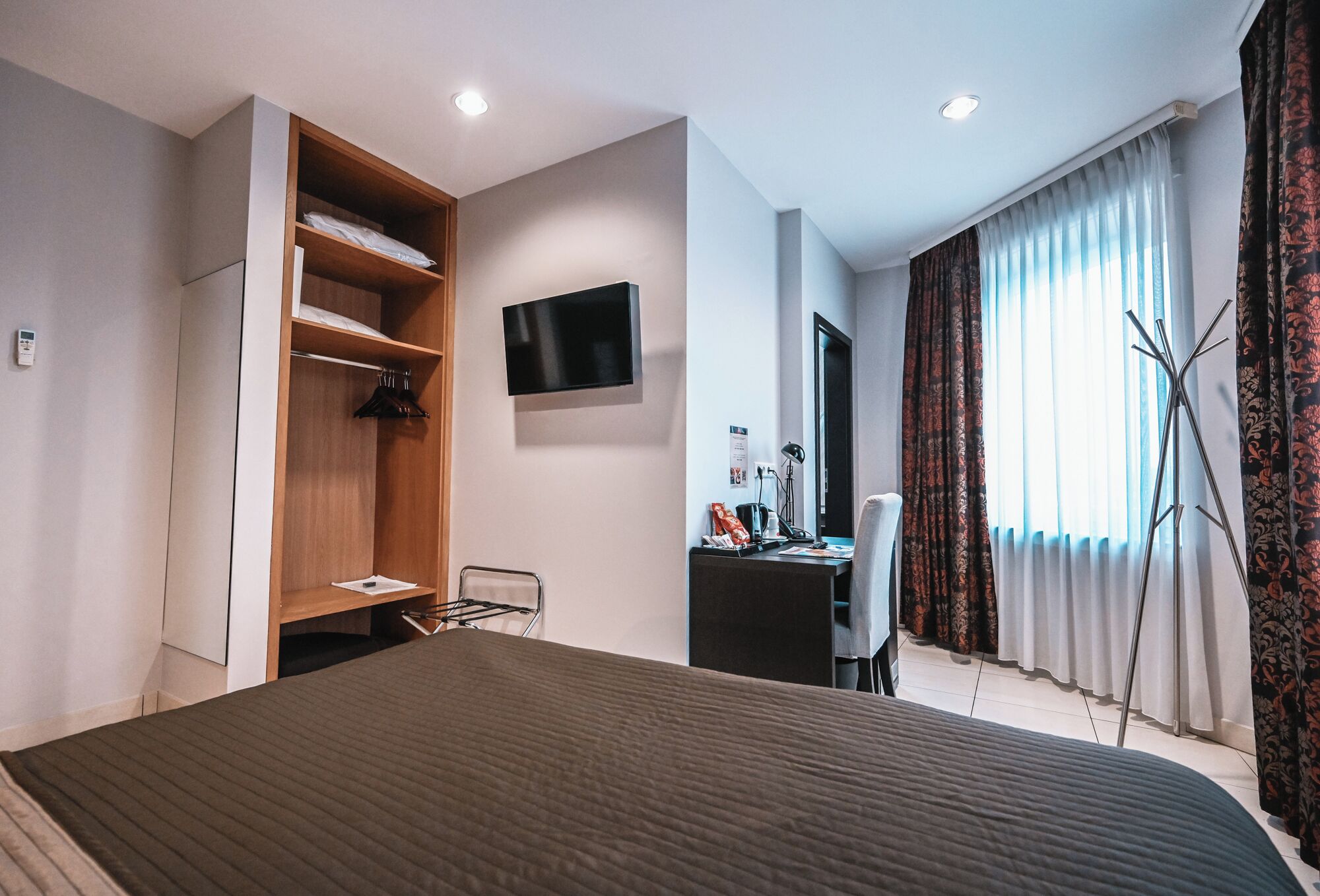 A modern hotel room with a large bed, a wooden wardrobe, a desk with a chair, a flat-screen TV, and a large window with curtains.