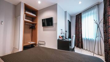 A modern hotel room with a large bed, a wooden wardrobe, a desk with a chair, a flat-screen TV, and a large window with curtains.