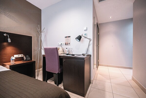 A hotel room with a bed, a desk with a chair, a desk lamp, a telephone, and a small table with a water bottle and a glass.