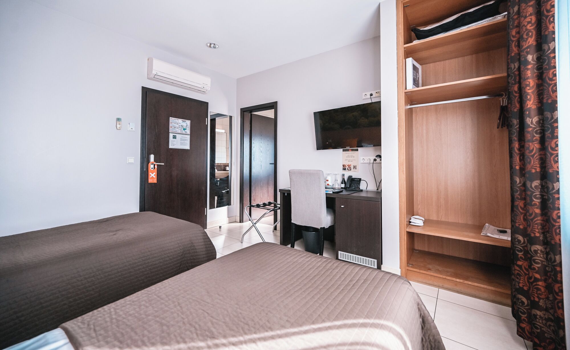 Habitación de hotel con dos camas, un escritorio con silla, armario, televisor y baño visible a través de una puerta abierta.