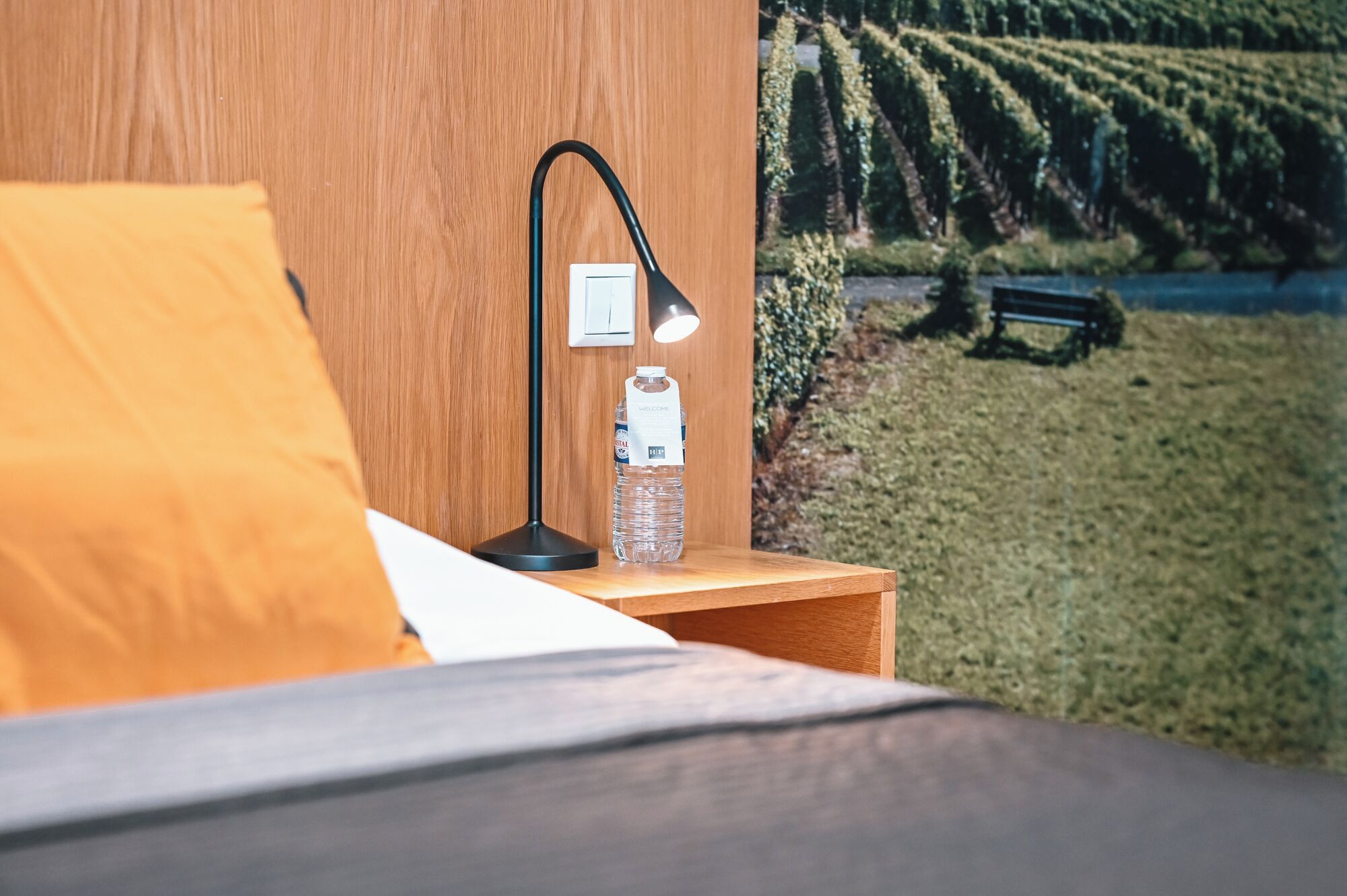 A neatly made bed with a yellow pillow, a black desk lamp, and a water bottle on a wooden nightstand.