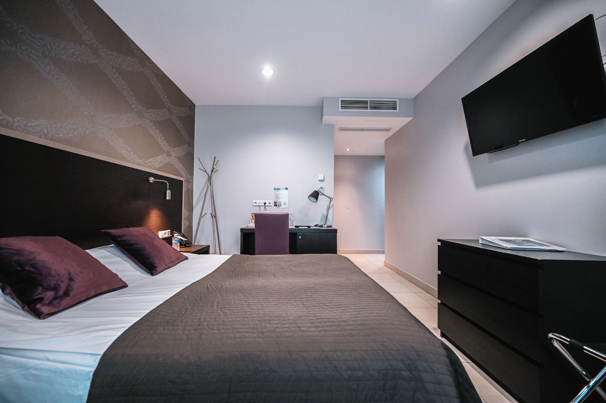 Quarto de hotel moderno com uma cama grande, uma TV de tela plana na parede, uma escrivaninha com luminária e uma cadeira.