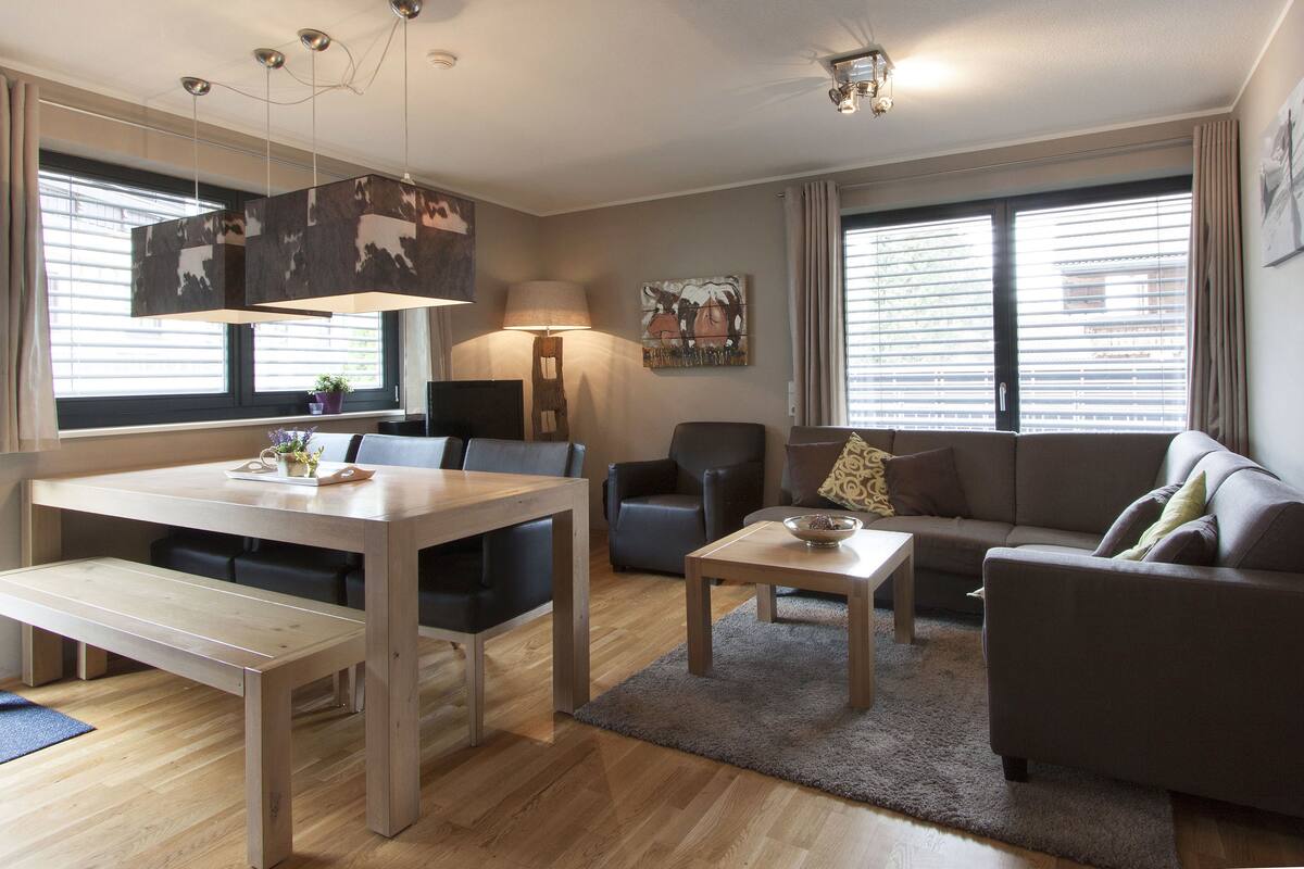 A modern living room with a dining table, sofa, and chairs. There is a lamp, a painting on the wall, and a window with blinds.