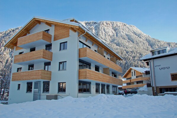 A multi-story building with wooden balconies, a ski slope in the background, and a clear sky.
