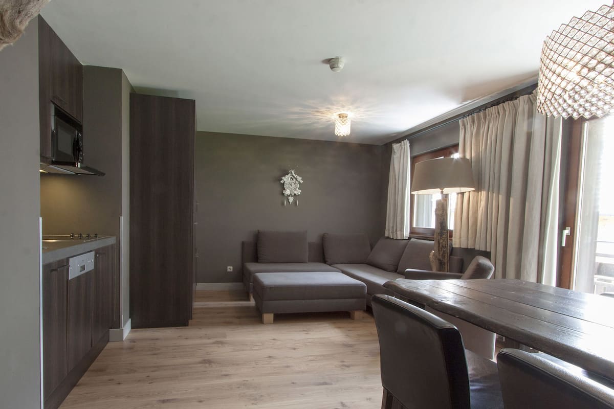 A modern kitchen with dark wood cabinets and a built-in stove. There is a gray sofa and a wooden dining table with chairs. A decorative wall piece hangs above the sofa.