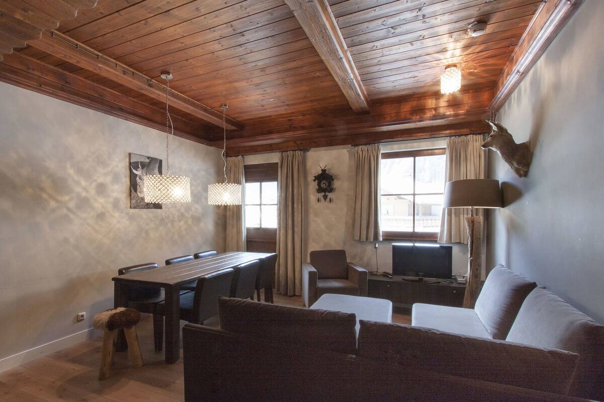 A cozy room with a wooden ceiling, a dining table with chairs, a sofa, a TV, and a lamp.