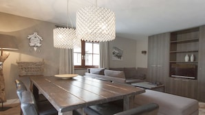 A modern dining area with a wooden table, chairs, and a chandelier. There is a sofa and a television in the background. - Appart Laijola (Gaschurn)