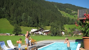 A swimming pool with people around it, surrounded by houses and a forested hillside. - Appart Laijola (Gaschurn)