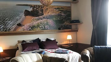 A cozy hotel room with a bed, a leather armchair, a round coffee table, and a large painting of a coastal landscape.