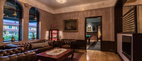 A spacious living room with a leather sofa, a wooden coffee table, and a chandelier. There are large windows with arched tops, a painting on the wall, and a billiards table in the background.