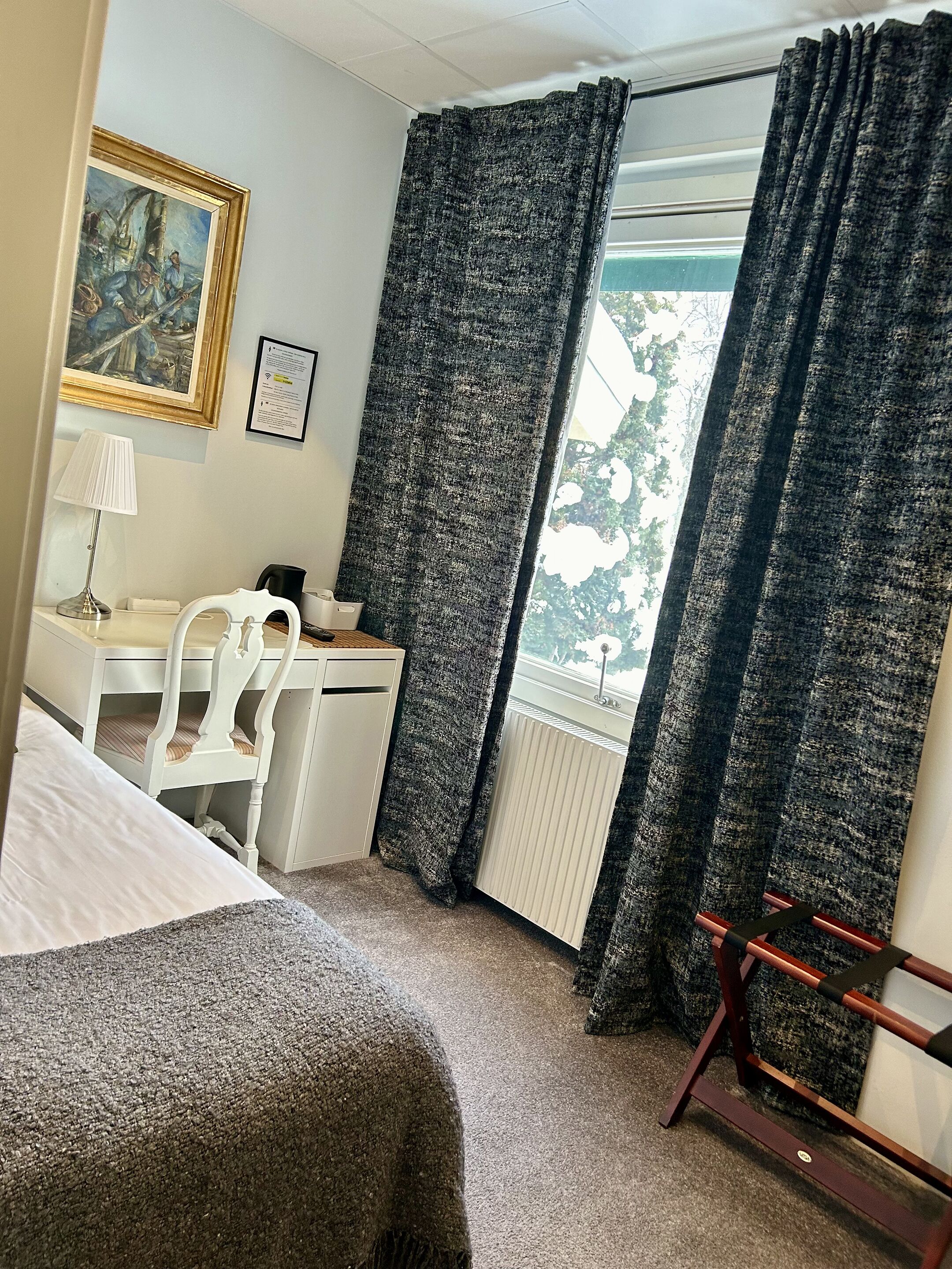 A hotel room with a bed, a desk, a chair, a radiator, and a window with curtains.