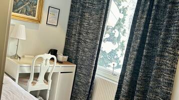 A hotel room with a bed, a desk, a chair, a radiator, and a window with curtains.
