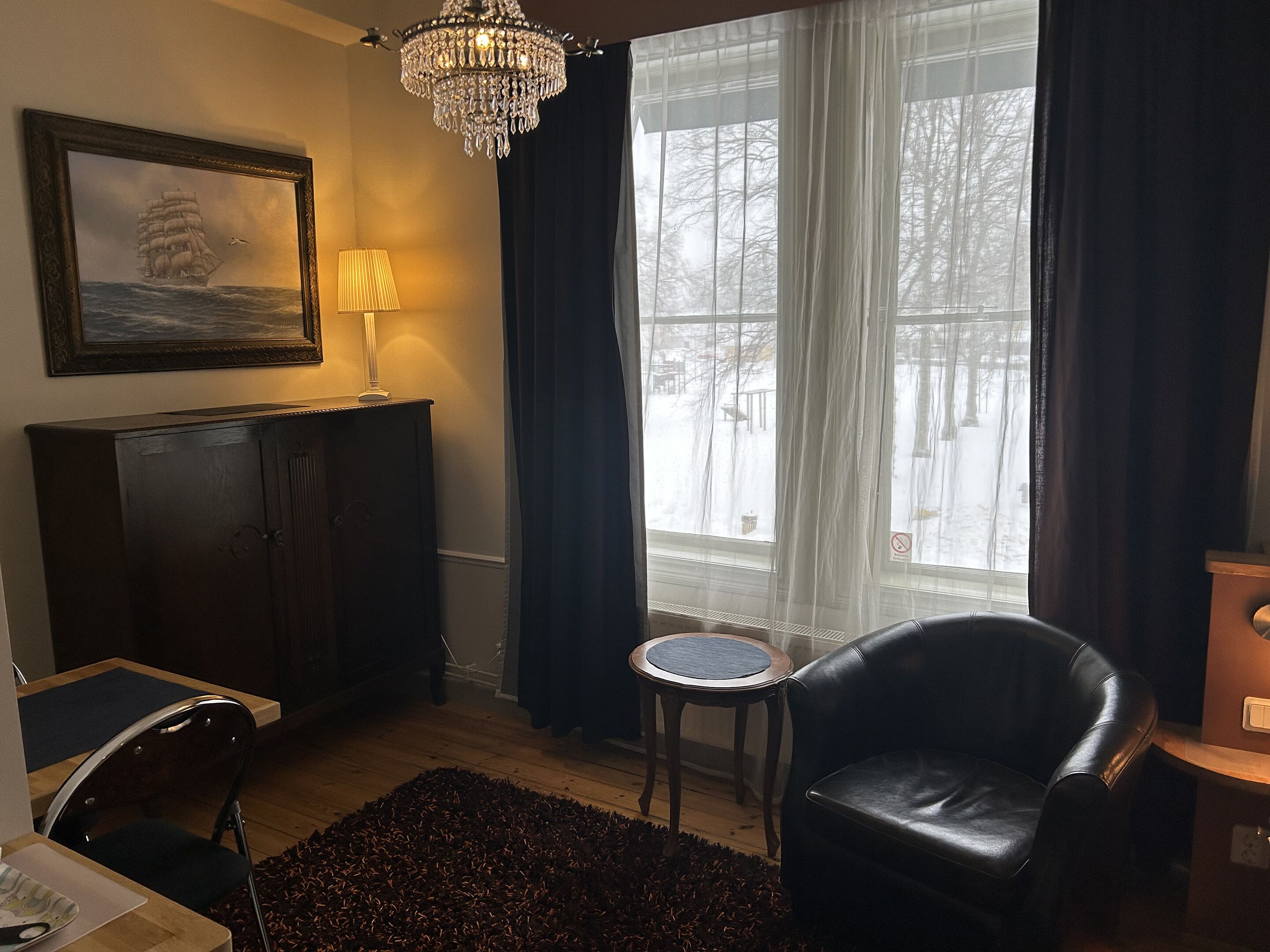 Ett mysigt rum med en mörk läderfåtölj, ett litet runt bord, en lampa, en inramad tavla som föreställer ett skepp och ett stort fönster med gardiner.