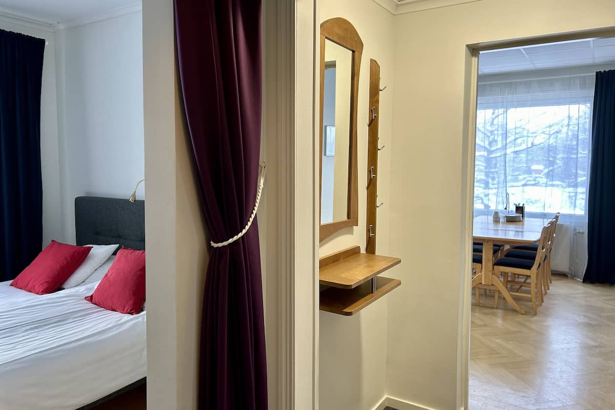 A hotel room with a bed, a mirror, a wooden side table, and a view of a dining area.