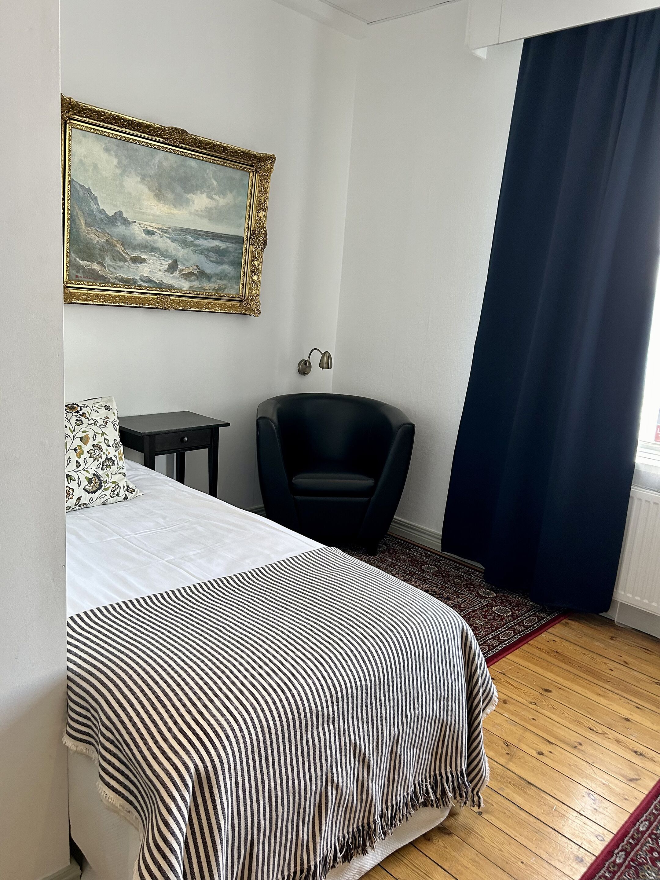 Un dormitorio con una cama, una silla, una mesita, una lámpara y un cuadro en la pared.