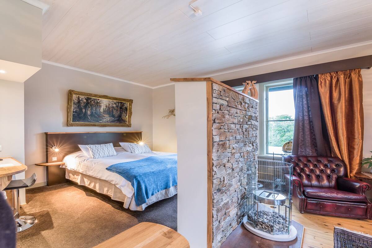 A bedroom with a bed, bedside table, and a nightstand. There is a window with curtains and a view of greenery outside.