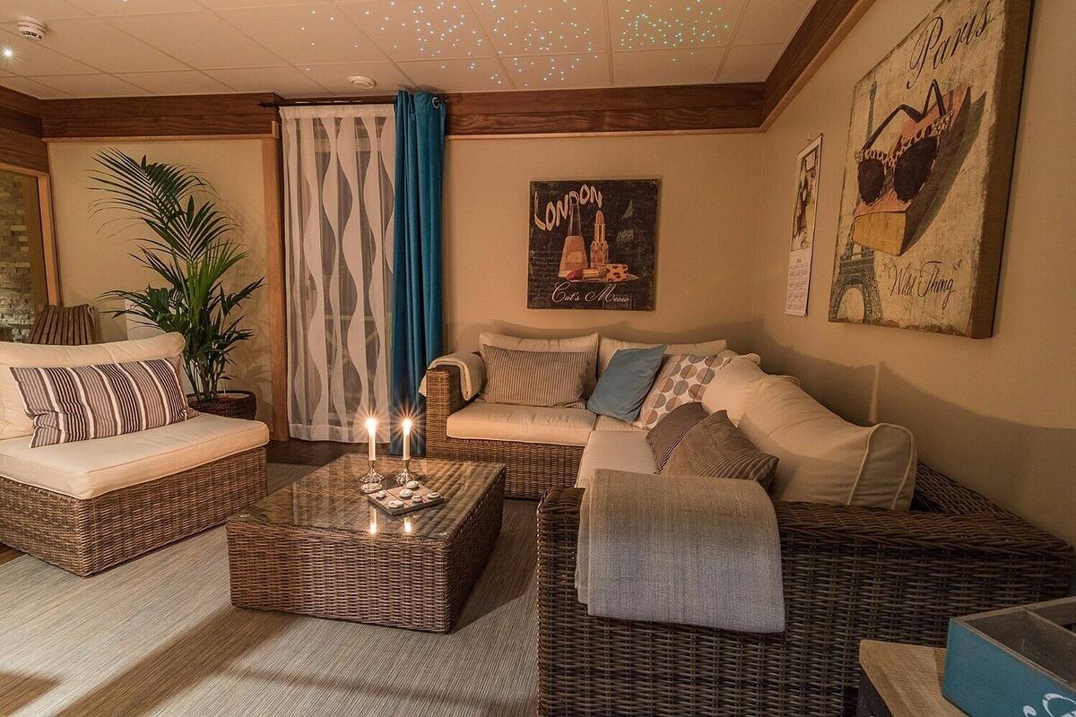 A cozy living room with a sofa, a coffee table, and a wicker chair. There are two potted plants and a wall with a poster.