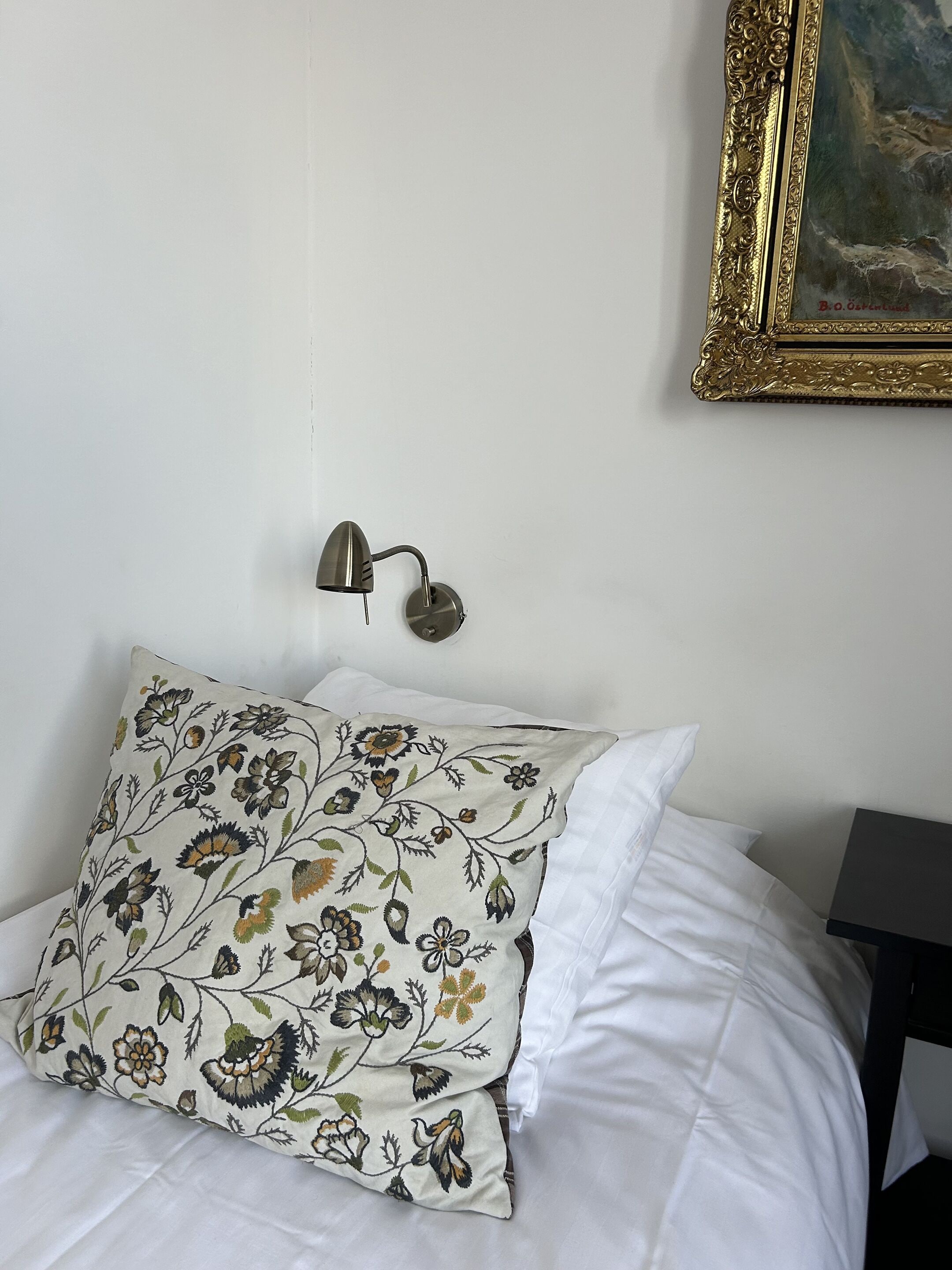 A bed with white bedding, a floral pillow, a wall-mounted lamp, and a framed painting on the wall.