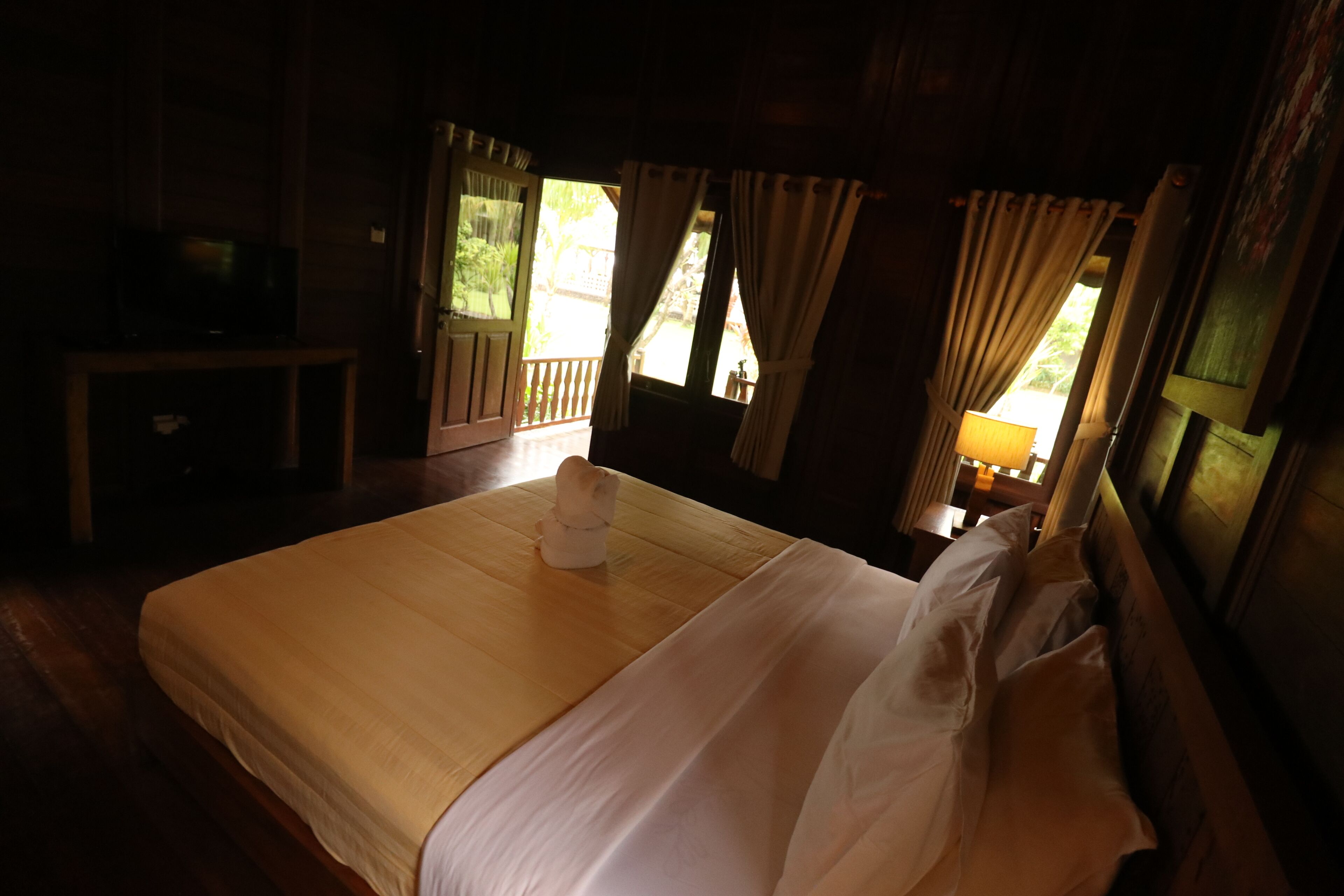 A neatly made bed with a white duvet and pillows, a wooden headboard, a small bedside table with a lamp, and a view of greenery through a window with curtains.