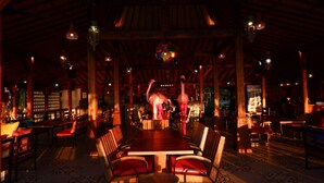 A dimly lit restaurant with wooden beams and hanging lanterns. Tables are set with red chairs, and a large elephant statue is visible in the center. - Mina Tanjung Hotel (Tanjung)