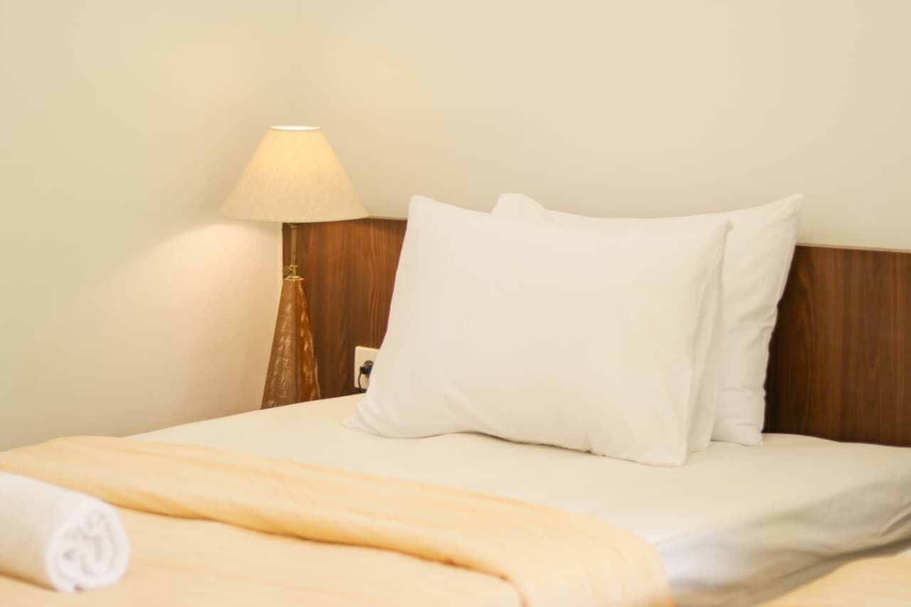 A neatly made bed with white linens, a rolled towel, and a bedside lamp.