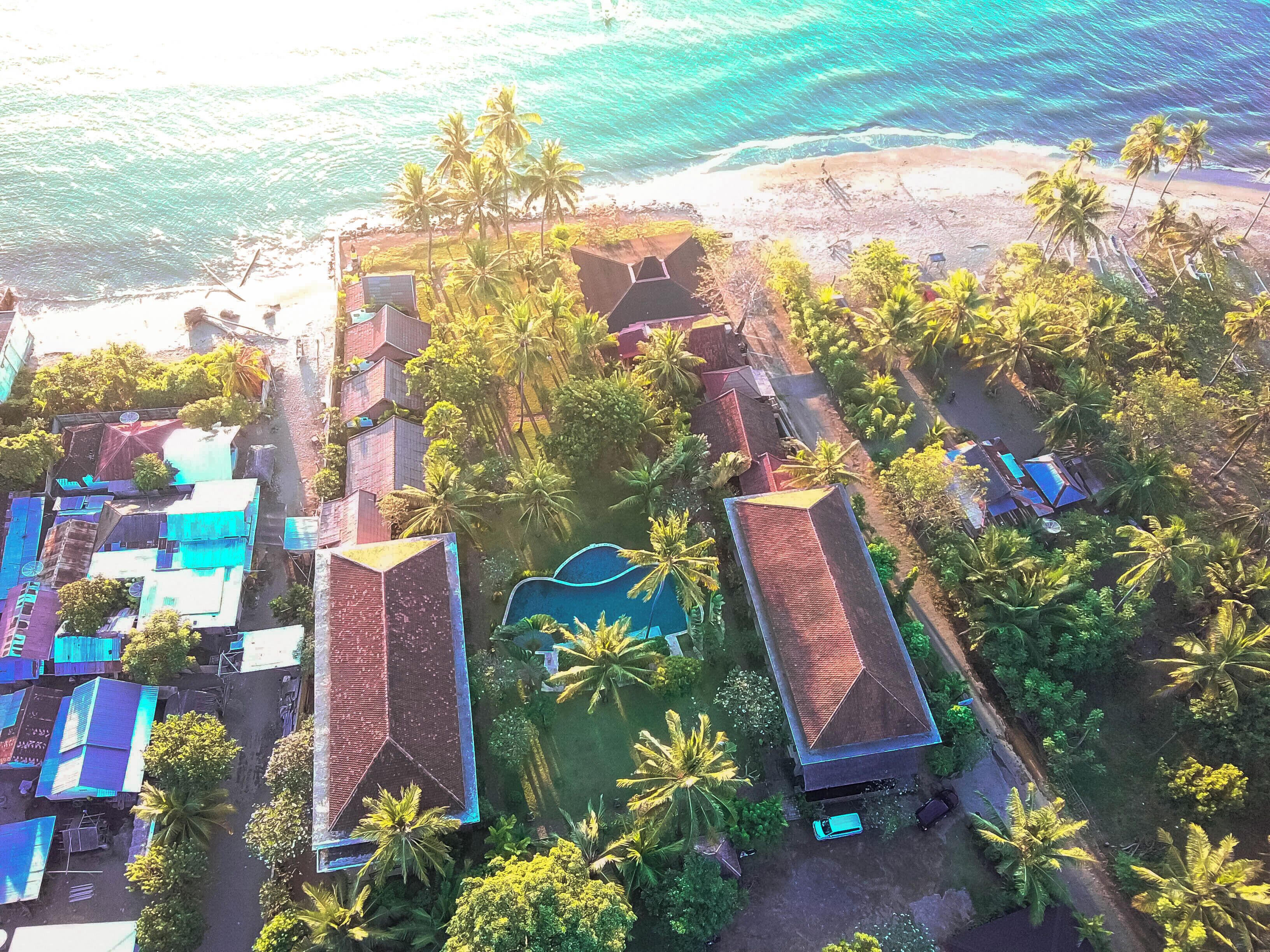 Luftaufnahme eines Küstenresorts mit mehreren Gebäuden, darunter ein Poolbereich, umgeben von Palmen und direktem Strandzugang.