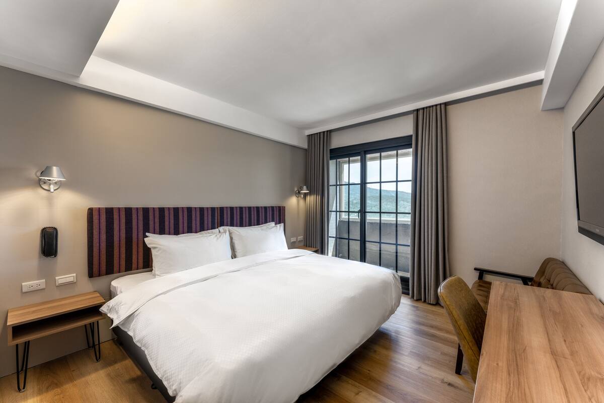 A modern hotel room with a large bed, a wooden desk, a seating area, and a large window with curtains.