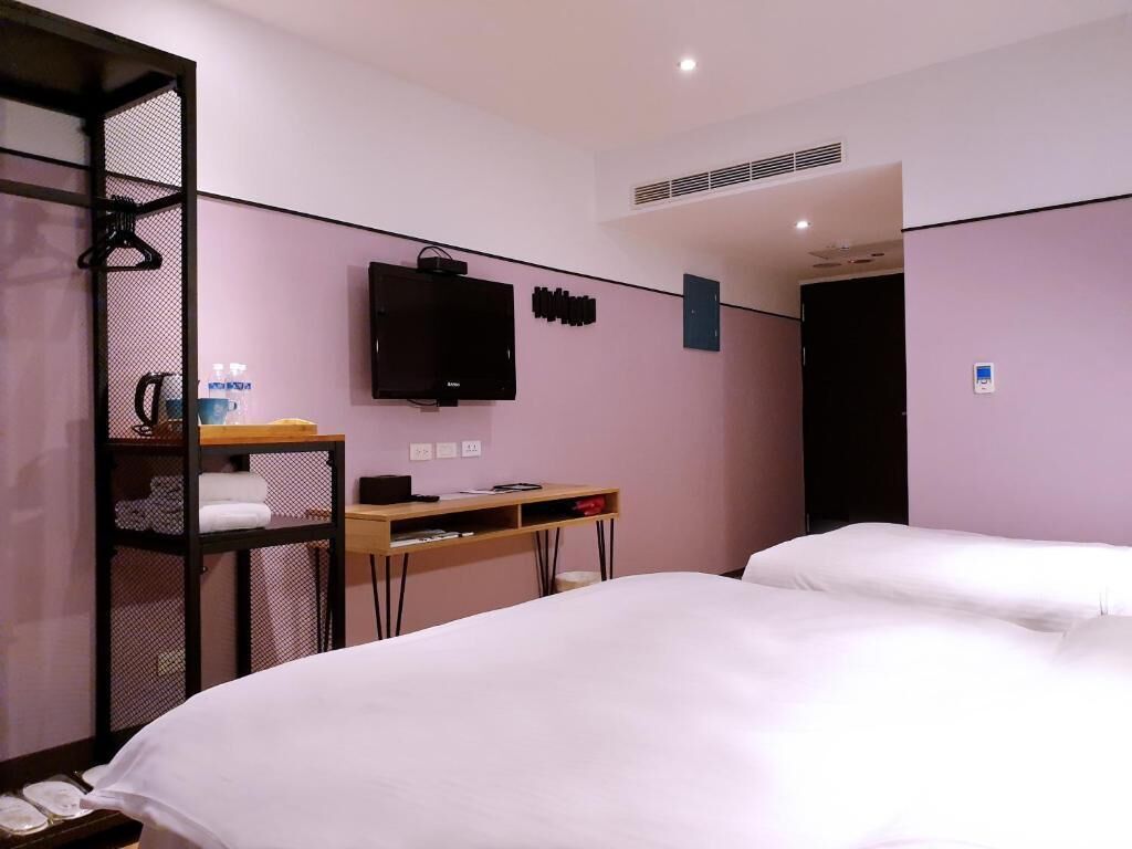A hotel room with two beds, a desk, a TV, and a shelf with towels.