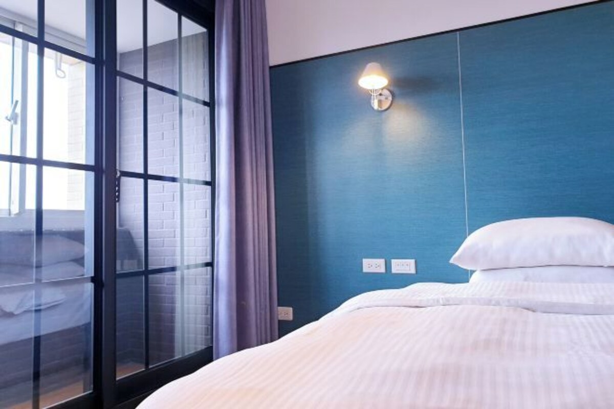 Una habitación de hotel con cama, ventana con cortinas, una lámpara de pared y un enchufe.