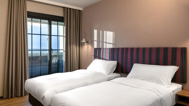 Habitación de hotel con dos camas, un ventanal grande con cortinas, cabecera a rayas y una mesita de noche.