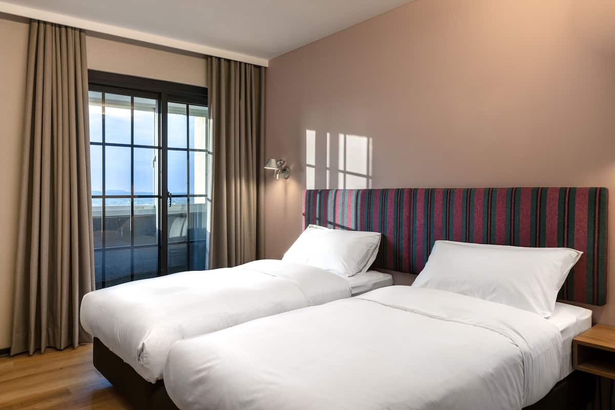 A hotel room with two beds, a large window with curtains, a striped headboard, and a nightstand.
