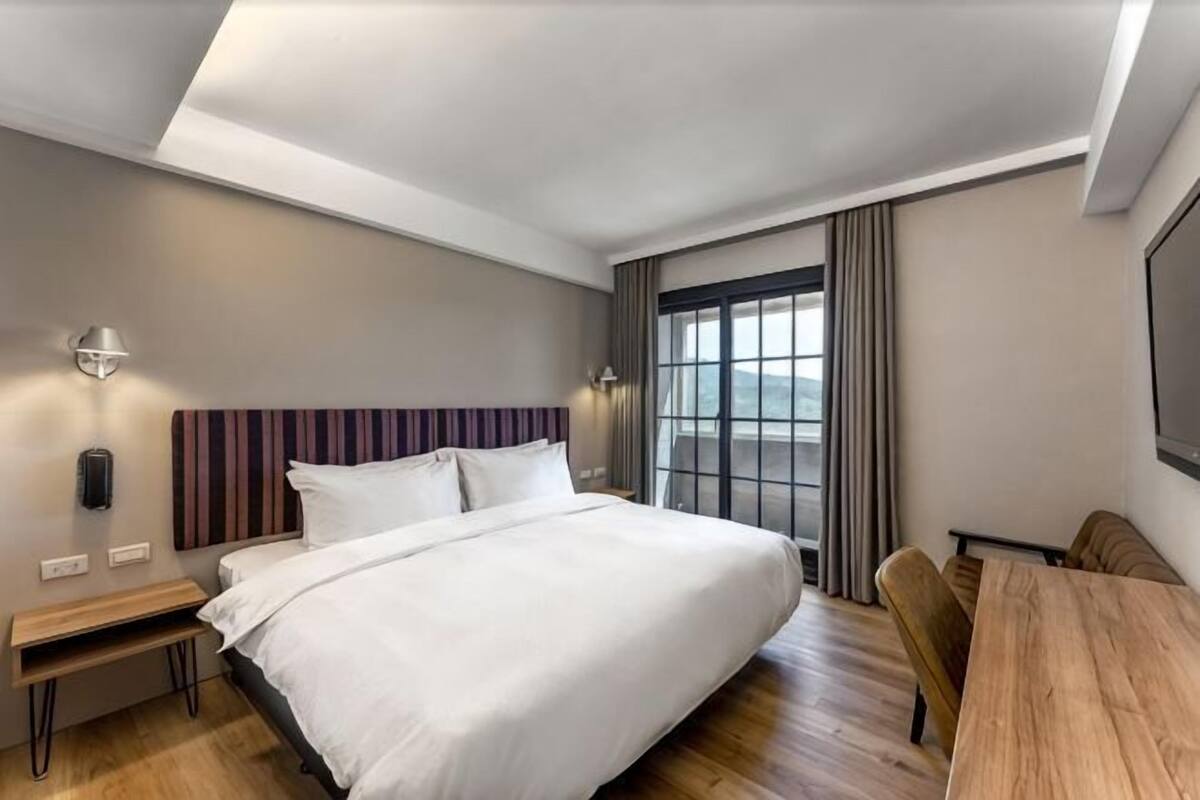 A modern hotel room with a large bed, bedside tables, a desk, and a view of the outdoors.