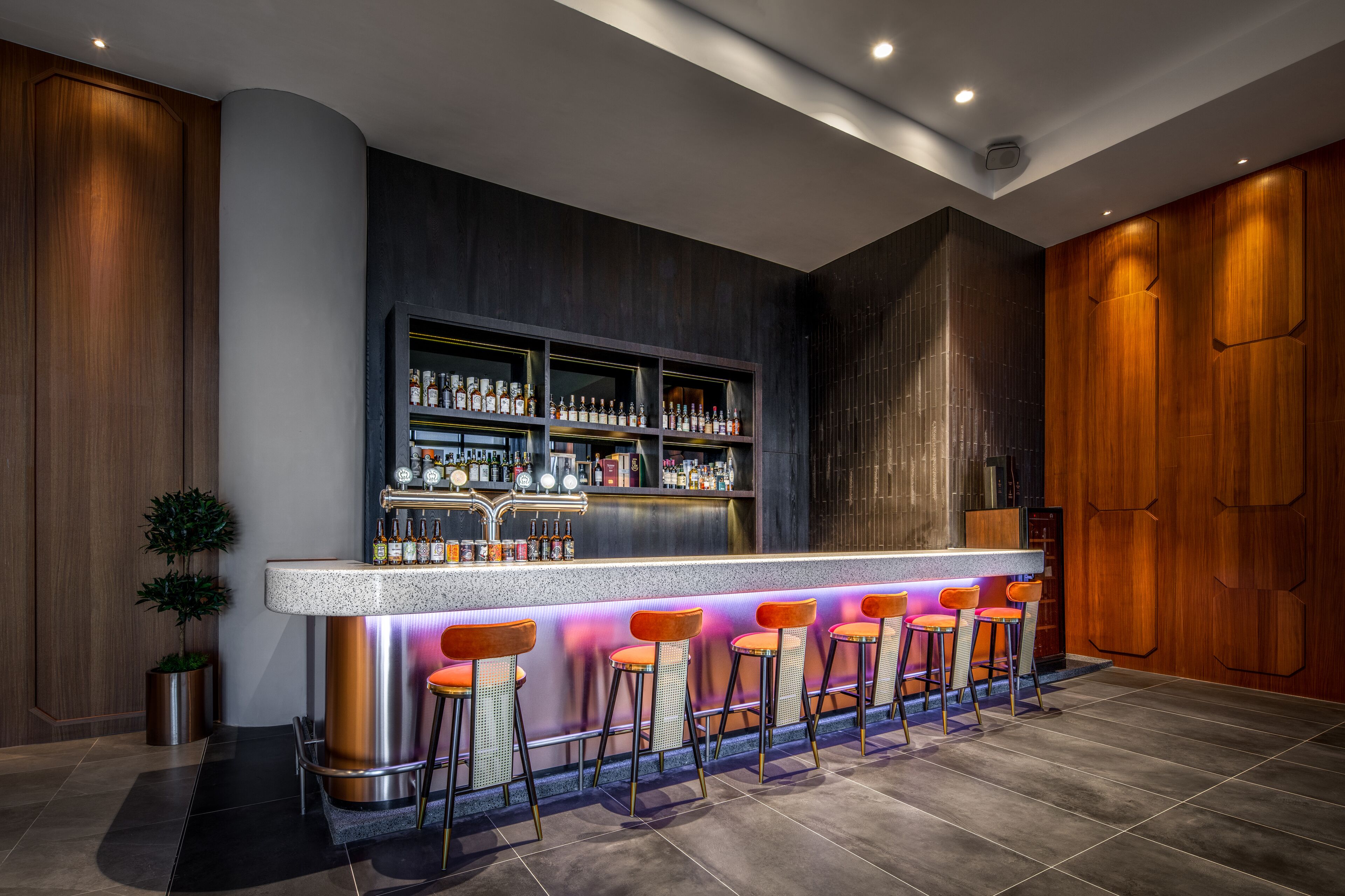 A modern bar area with a bar counter, multiple stools, and a well-stocked shelf with bottles.
