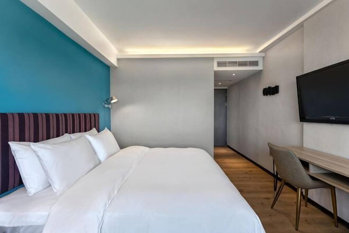 A hotel room with a bed, a desk, a chair, a TV, and a hallway leading to another room.