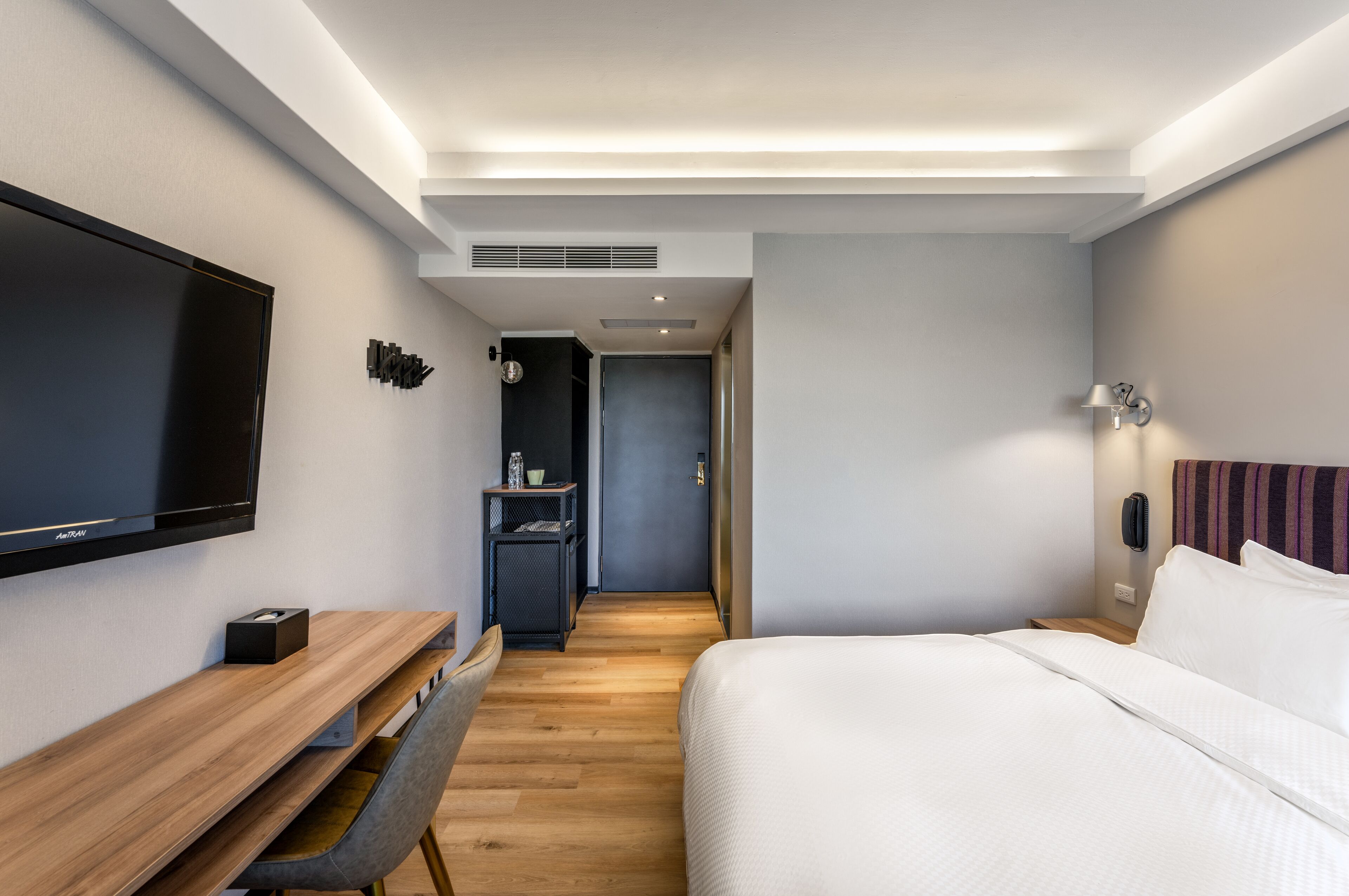 Une chambre d’hôtel moderne dotée d’une télévision à écran plat, d’un bureau en bois, d’un lit avec du linge de lit blanc et d’un petit coin salon.