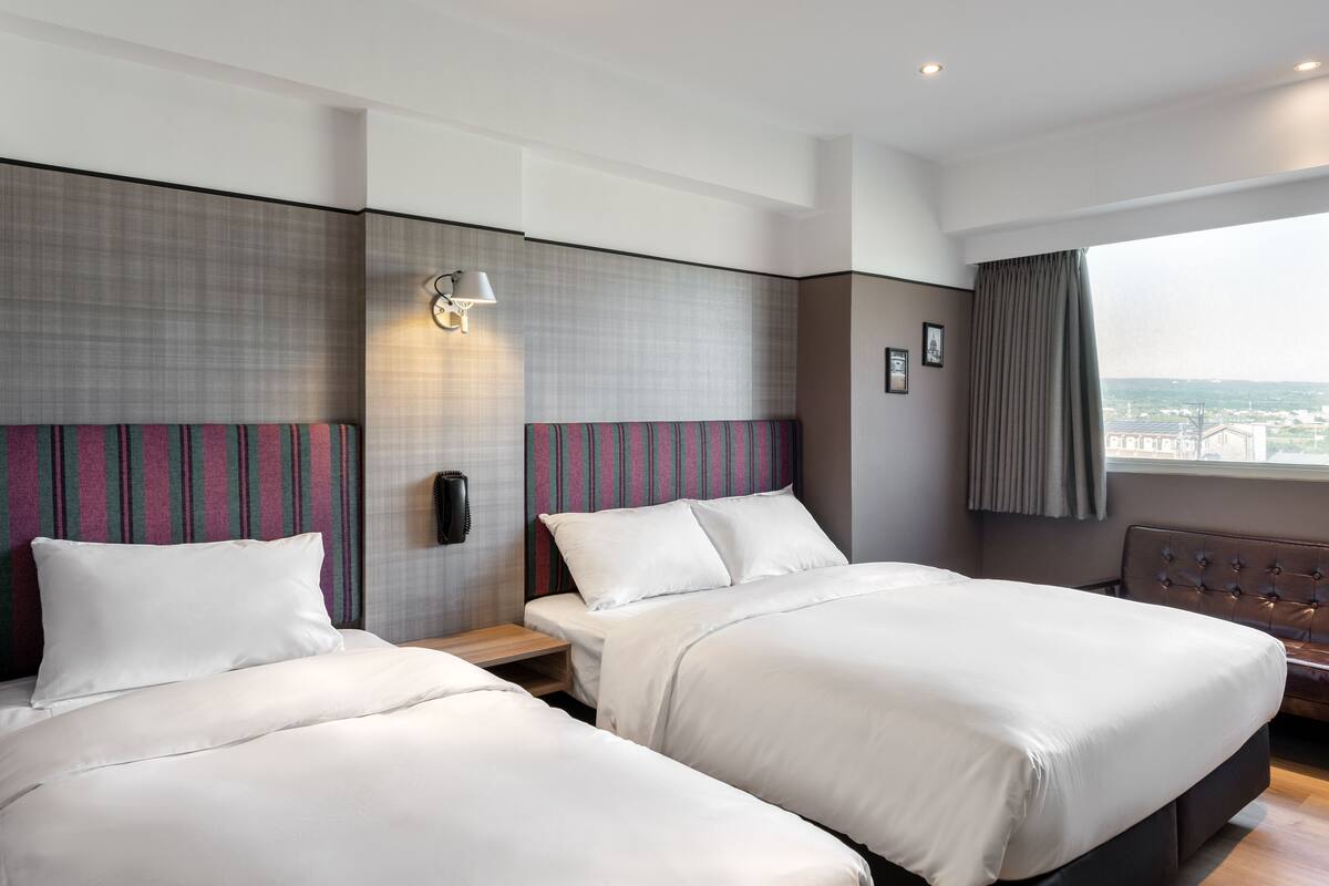 A hotel room with two beds, a large window, and a city view.