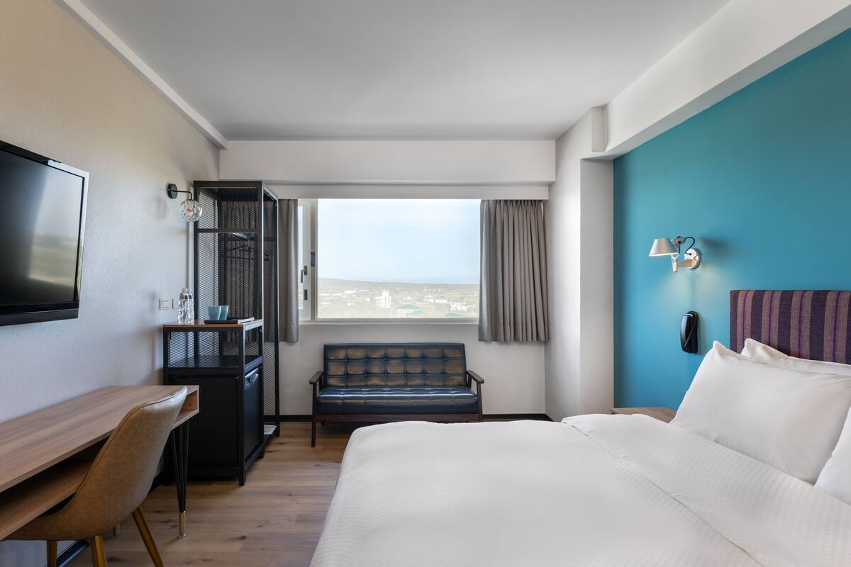 A hotel room with a bed, a desk, a chair, a TV, a window with a view, and a phone on the wall.