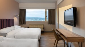 Habitación de hotel con dos camas, un escritorio, una silla, un televisor y una ventana con vista al mar.